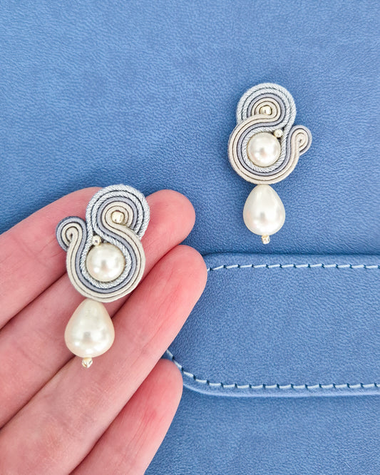 A pair of soutache earrings with white pearls and silver-toned braided details, shown with one earring on a blue textured surface and the other held on a person's fingers.