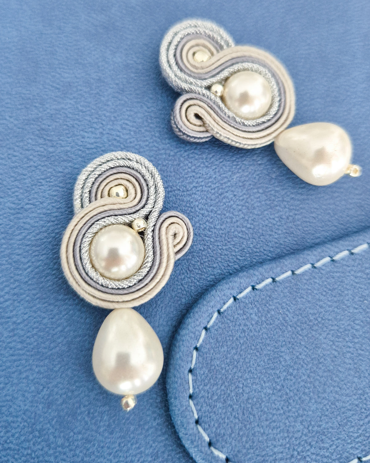 A pair of handmade soutache earrings featuring cream and silver braided cord, a central white pearl, and a dangling teardrop-shaped white pearl, displayed on a textured blue surface.
